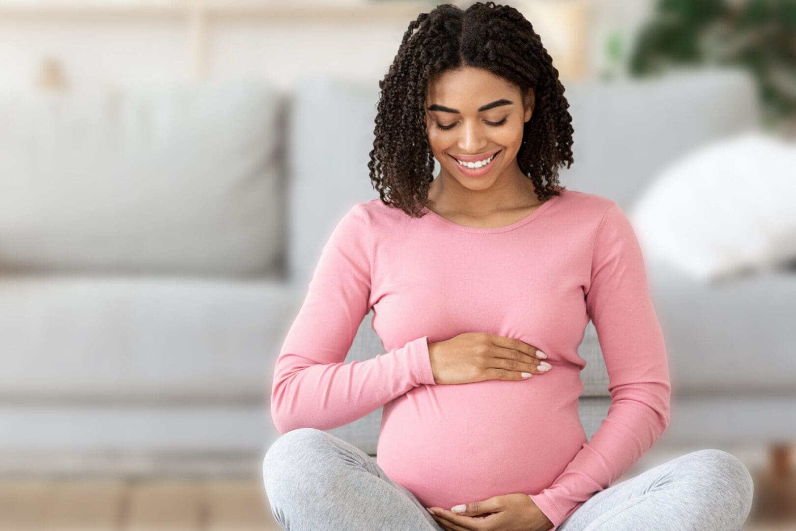 Beautiful happy pregnant black woman touching her tummy, sitting on sport mat at home, free space
