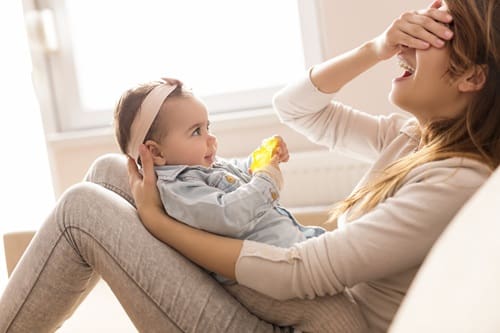 Young mother holding her baby girl in her arms sitting on a living room couch next to a window laughing and playing