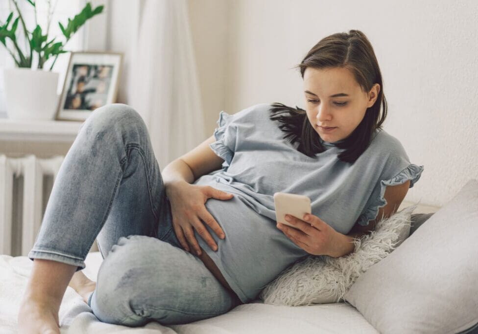 Pregnant woman using phone on bed.