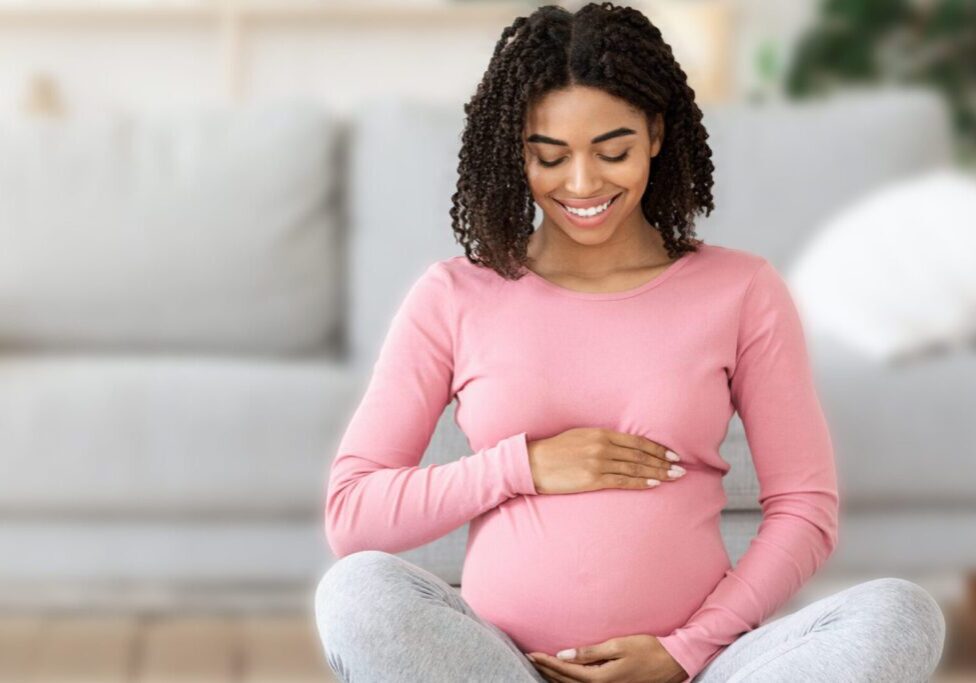 Beautiful happy pregnant black woman touching her tummy, sitting on sport mat at home, free space